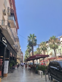 A lively street scene with people walking and sitting at outdoor tables under a maroon awning that reads 'Mon Chéri'. Tall palm trees line the street alongside a row of pastel-colored buildings. On the left, a store displays cushions, and a large black vehicle is parked on the right. The sky is clear and blue.