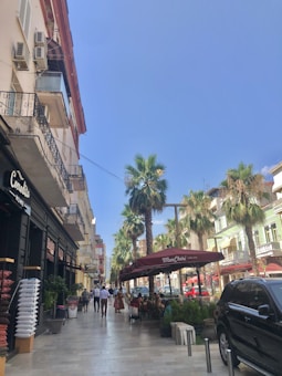 A lively street scene with people walking and sitting at outdoor tables under a maroon awning that reads 'Mon Ch&eacute;ri'. Tall palm trees line the street alongside a row of pastel-colored buildings. On the left, a store displays cushions, and a large black vehicle is parked on the right. The sky is clear and blue.
