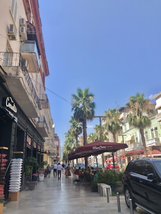A lively street scene with people walking and sitting at outdoor tables under a maroon awning that reads 'Mon Ch&eacute;ri'. Tall palm trees line the street alongside a row of pastel-colored buildings. On the left, a store displays cushions, and a large black vehicle is parked on the right. The sky is clear and blue.