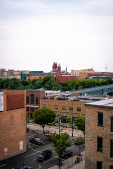 A cityscape featuring a mix of modern and historic buildings. The foreground includes Brown's Race Market, identifiable by its signage, with a few trees and parked cars lining the street. The background shows industrial structures and a bridge under a cloudy sky.
