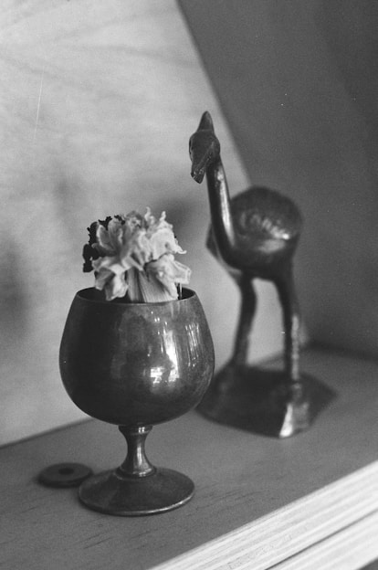 A vintage, metallic goblet holds a wilted flower on a wooden surface. In the background, a metal figurine of a bird stands, showcasing an antiquated, artistic style. The overall composition exudes a nostalgic, timeless quality.