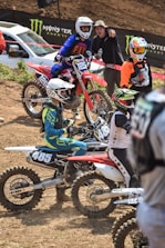 Several motocross riders are gathered on a dirt track, with dirt bikes marked with numbers 125 and 485. They are dressed in colorful racing gear and helmets. One person, not in racing gear, is standing and appears to be talking or cheering. A white vehicle and Monster Energy brand banners are visible in the background.