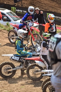 Several motocross riders are gathered on a dirt track, with dirt bikes marked with numbers 125 and 485. They are dressed in colorful racing gear and helmets. One person, not in racing gear, is standing and appears to be talking or cheering. A white vehicle and Monster Energy brand banners are visible in the background.