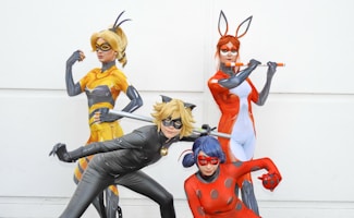 Four individuals dressed in colorful superhero costumes striking dynamic poses. Each costume is distinctive, featuring masks and thematic colors, such as yellow, black, orange, and red, with various accessories and props.