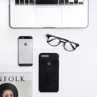 A flat lay composition on a white surface includes a silver laptop, a smartphone with a silver body, a black smartphone, a pair of black-rimmed glasses, and a magazine partially showing the title 'FOLK'.