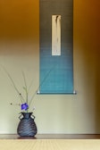 A traditional Japanese room featuring a woven basket with an arrangement of delicate purple and green flowers placed on a tatami mat. Behind it is a scroll hanging on the wall with subtle artwork and calligraphy, against a gradient background transitioning from dark to light blue.