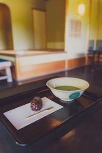 A traditional Japanese tea room with a focus on a black tray containing a bowl of green tea and a sweet treat on a piece of paper. The room features minimalistic wooden furniture and soft lighting, creating a serene and calm atmosphere.