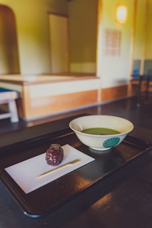A traditional Japanese tea room with a focus on a black tray containing a bowl of green tea and a sweet treat on a piece of paper. The room features minimalistic wooden furniture and soft lighting, creating a serene and calm atmosphere.