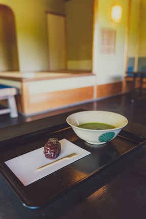 A traditional Japanese tea room with a focus on a black tray containing a bowl of green tea and a sweet treat on a piece of paper. The room features minimalistic wooden furniture and soft lighting, creating a serene and calm atmosphere.