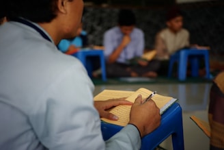 A person is seated at a small blue table, writing in a notebook filled with text in a language that resembles Arabic script. In the background, several other individuals are seated on similar blue stools, likely engaged in reading or studying. The setting suggests a classroom or study group environment.