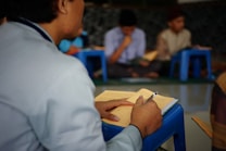 A person is seated at a small blue table, writing in a notebook filled with text in a language that resembles Arabic script. In the background, several other individuals are seated on similar blue stools, likely engaged in reading or studying. The setting suggests a classroom or study group environment.
