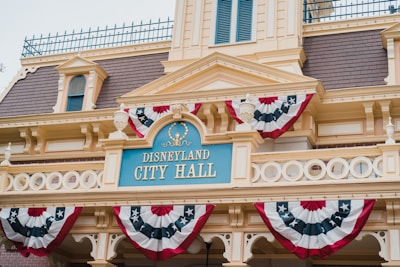 A building with an ornate facade featuring classic architectural elements like columns and decorative moldings. It prominently displays a sign reading 'Disneyland City Hall' in light blue with gold detailing. Above the sign, there are red, white, and blue patriotic bunting draped along the balcony.