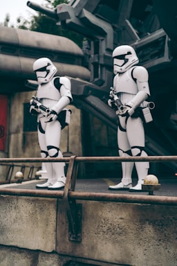 Two individuals dressed as stormtroopers stand on a platform in front of a sci-fi or futuristic backdrop. Both figures are in white armor, holding blaster guns, with a metallic railing in the foreground and industrial structures in the background.
