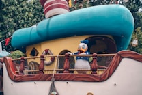 A whimsical boat structure features a cartoon character in a sailor outfit, standing on the deck. The boat has bright colors with a predominantly teal and beige color scheme, incorporating playful elements like lifebuoys and decorative railing. The background is lush with greenery.
