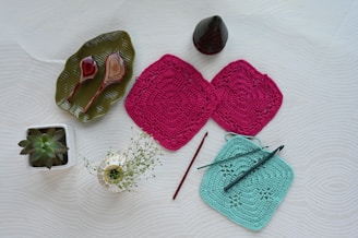 A flat lay arrangement featuring two red crochet squares, a teal crochet square with a crochet hook placed on top, a red crochet hook, a green potted succulent plant, a small vase with white flowers, a green leaf-shaped plate holding two carved wooden spoons, and a small cone-shaped object.