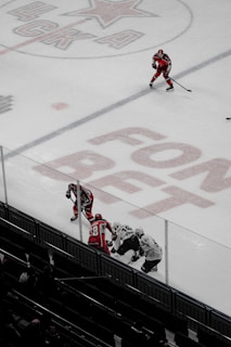 A hockey game on an ice rink with players wearing red and white uniforms. The number 48 and advertisements are visible on the ice. Three players engage closely near the boards, while another skater positions further up the rink, holding a hockey stick.