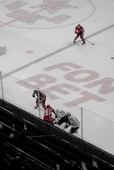 A hockey game on an ice rink with players wearing red and white uniforms. The number 48 and advertisements are visible on the ice. Three players engage closely near the boards, while another skater positions further up the rink, holding a hockey stick.