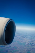 A jet engine is prominently featured on the left side with a view of the landscape and horizon below. The sky is clear and blue, stretching endlessly.