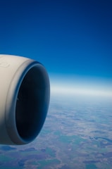 A jet engine is prominently featured on the left side with a view of the landscape and horizon below. The sky is clear and blue, stretching endlessly.