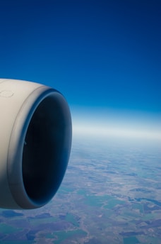 A jet engine is prominently featured on the left side with a view of the landscape and horizon below. The sky is clear and blue, stretching endlessly.