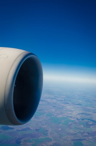 A jet engine is prominently featured on the left side with a view of the landscape and horizon below. The sky is clear and blue, stretching endlessly.