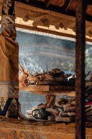 An arrangement of traditional offerings sits on an ornately decorated wooden stand. Incense sticks are lit and smoke is gently rising. The stand is draped with rich fabrics featuring intricate patterns. Natural light filters through, casting soft shadows and highlighting the textures of the materials.