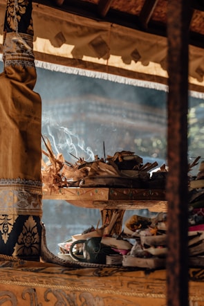 An arrangement of traditional offerings sits on an ornately decorated wooden stand. Incense sticks are lit and smoke is gently rising. The stand is draped with rich fabrics featuring intricate patterns. Natural light filters through, casting soft shadows and highlighting the textures of the materials.