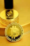 A stack of shiny, gold-like cryptocurrency coins featuring the logos of Ethereum and Bitcoin. The coins have a metallic sheen, with the Ethereum coin displaying a triangular symbol and Bitcoin displaying a 'B' with two vertical lines running through it.