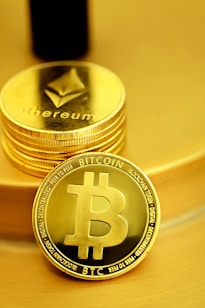 A stack of shiny, gold-like cryptocurrency coins featuring the logos of Ethereum and Bitcoin. The coins have a metallic sheen, with the Ethereum coin displaying a triangular symbol and Bitcoin displaying a 'B' with two vertical lines running through it.
