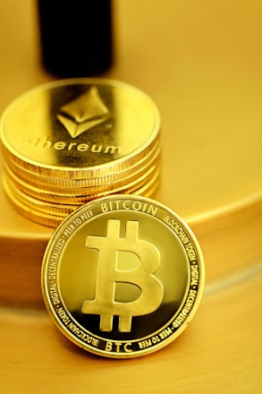 A stack of shiny, gold-like cryptocurrency coins featuring the logos of Ethereum and Bitcoin. The coins have a metallic sheen, with the Ethereum coin displaying a triangular symbol and Bitcoin displaying a 'B' with two vertical lines running through it.