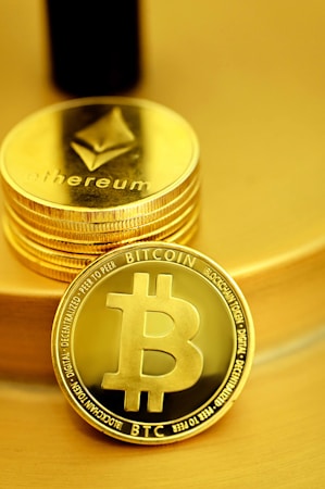 A stack of shiny, gold-like cryptocurrency coins featuring the logos of Ethereum and Bitcoin. The coins have a metallic sheen, with the Ethereum coin displaying a triangular symbol and Bitcoin displaying a 'B' with two vertical lines running through it.