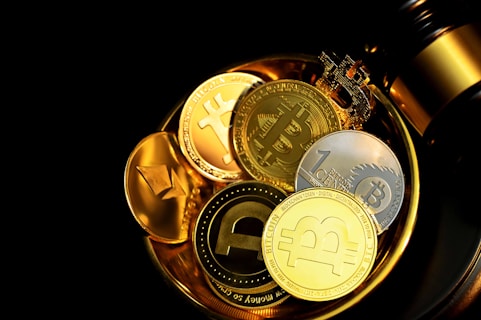 A collection of various cryptocurrency coins displayed against a dark background, with a blend of golden, silver, and bronze tones. Visible coins include representations of Bitcoin, Ethereum, and other crypto symbols, placed in a shiny metallic dish or cup. A gavel is partially visible, suggesting themes of legality or auction.