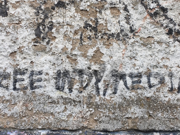 A roughly textured, weathered wall features partially faded, black graffiti with the lettering 'FREE' followed by what appears to be 'FREEDOM'. The wall's surface is uneven, with chipped paint revealing underlying layers.