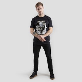 A person stands against a plain gray background wearing a black t-shirt with white and gold graphics. The design includes text and intricate patterns. The individual is dressed in black pants and black shoes with brown soles. Tattoos are visible on their forearms, and they have a neutral expression with arms loosely crossed.