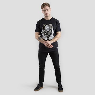 A person stands against a plain gray background wearing a black t-shirt with white and gold graphics. The design includes text and intricate patterns. The individual is dressed in black pants and black shoes with brown soles. Tattoos are visible on their forearms, and they have a neutral expression with arms loosely crossed.