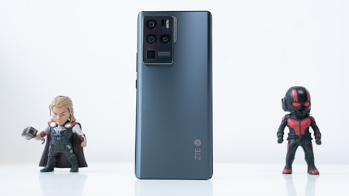 A black ZTE smartphone is placed on a white surface with two action figures on either side. The phone's back is facing the camera, showing its camera module. The action figure on the left is holding a hammer and wearing a cape, while the one on the right is dressed in a black and red suit with a helmet.