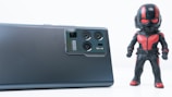 A sleek, modern smartphone is lying on a flat surface, showcasing its advanced camera module. Beside the smartphone stands a figurine of a superhero dressed in a black and red costume, wielding a helmet with bright red accents. The clean, white background accentuates the objects, creating a minimalist aesthetic.