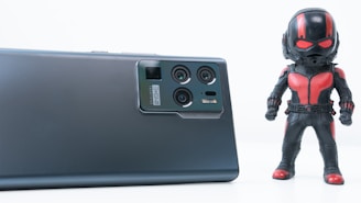 A sleek, modern smartphone is lying on a flat surface, showcasing its advanced camera module. Beside the smartphone stands a figurine of a superhero dressed in a black and red costume, wielding a helmet with bright red accents. The clean, white background accentuates the objects, creating a minimalist aesthetic.