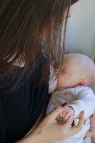 A woman is nursing a baby. The woman's long brown hair partially covers her face as she gazes down at the baby. The baby is close to her chest, wearing a white outfit, and is feeding. The woman's hand gently holds the baby's tiny hand.