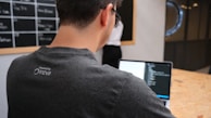 A man is working on a laptop, coding or programming, wearing a dark t-shirt with a logo on the back. In the background, a blackboard with visible text and a person standing near it are partially seen. The setting appears to be an office or workspace with a wooden table.