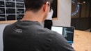 A man is working on a laptop, coding or programming, wearing a dark t-shirt with a logo on the back. In the background, a blackboard with visible text and a person standing near it are partially seen. The setting appears to be an office or workspace with a wooden table.