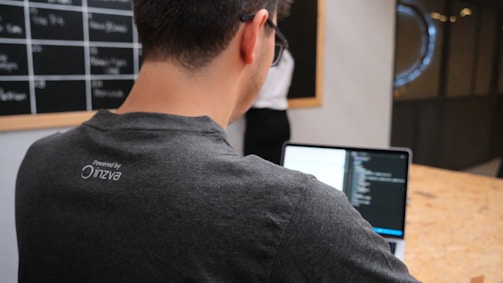 A man is working on a laptop, coding or programming, wearing a dark t-shirt with a logo on the back. In the background, a blackboard with visible text and a person standing near it are partially seen. The setting appears to be an office or workspace with a wooden table.