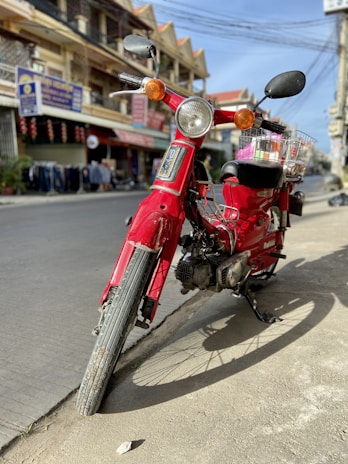 A red motorcycle is parked on a city street, tilted slightly on its kickstand. Surrounding the motorcycle are shops and urban buildings with various signs, indicating a lively daytime environment. The motorcycle has a basket mounted on the back, and the sun casts clear shadows on the pavement.