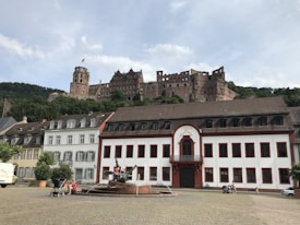 A historic castle sits atop a lush, green hillside, overlooking a plaza with neatly arranged old-style buildings. The castle is made of stone, featuring towers and ruinous walls indicative of its ancient origins. In the foreground, a decorative fountain with a statue is surrounded by a cobblestone square, where a few people can be seen walking and others are seated nearby.