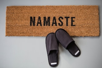 A doormat featuring the word 'Namaste' in black letters on a textured brown surface is placed on a light floor. Beside the mat, a pair of black slippers is positioned in a relaxed manner.