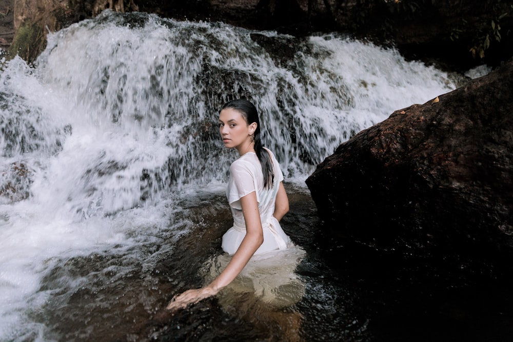 woman in white shirt and white shorts sitting on rock near water falls