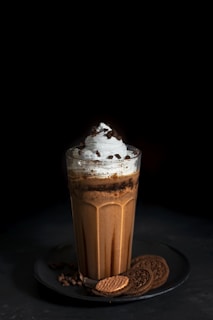 A tall glass of chocolate milkshake topped with whipped cream and chocolate chips, surrounded by chocolate cookies arranged on a dark plate. The background is dark, accentuating the dessert.