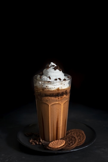 A tall glass of chocolate milkshake topped with whipped cream and chocolate chips, surrounded by chocolate cookies arranged on a dark plate. The background is dark, accentuating the dessert.