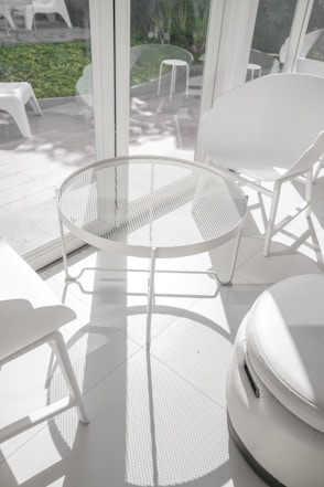 A sunlit room with modern white furniture, including a round mesh metal table, several white chairs, and stools. The room's large windows allow natural light to create intricate shadow patterns on the floor. Outside the windows, a garden with green plants and additional seating is visible.