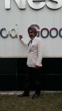 A man wearing a white suit and a turban is standing next to a large sign with letters. He has a beard and is posing with one hand resting on the sign. The ground he is standing on is covered with grass.